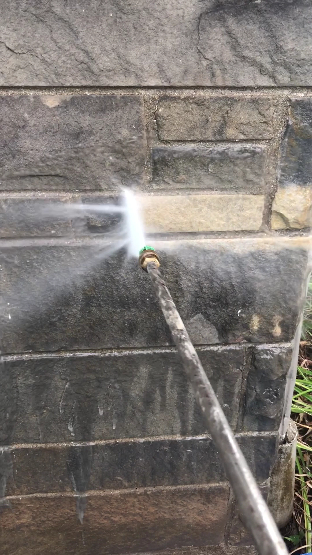 A pressure washing wand actively spraying and cleaning a grimy stone retaining wall
