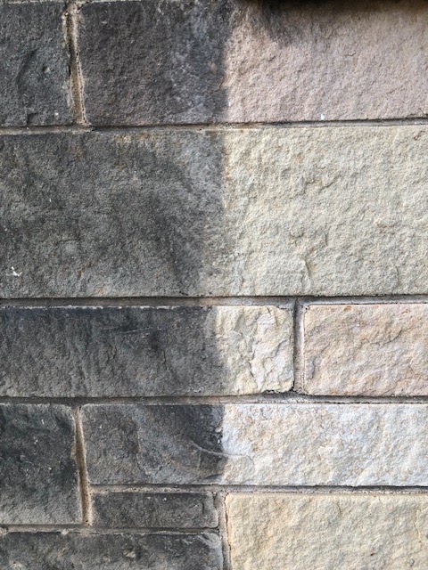 A stone wall, half still covered in dark grime and half freshly pressure washed to its natural light color, in one photo